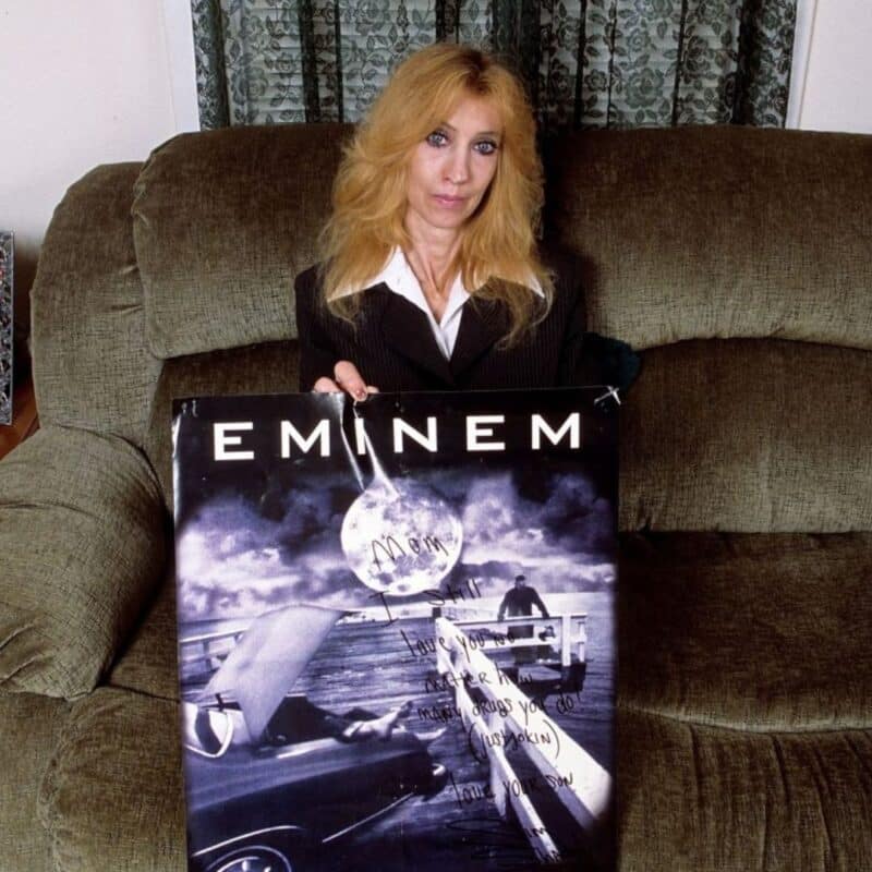 Eminem's mother, Debbie Mathers, sits on a couch holding a signed poster of Eminem