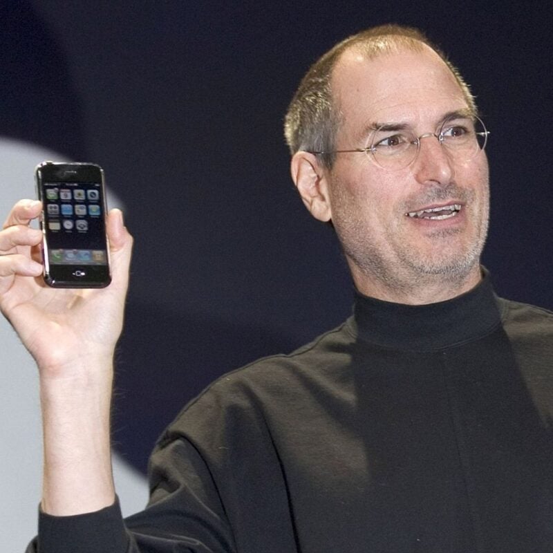 Steve Jobs holding an iPhone while speaking on stage during a presentation