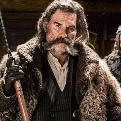 Kurt Russell in costume holding a rifle, wearing a fur coat and white shirt