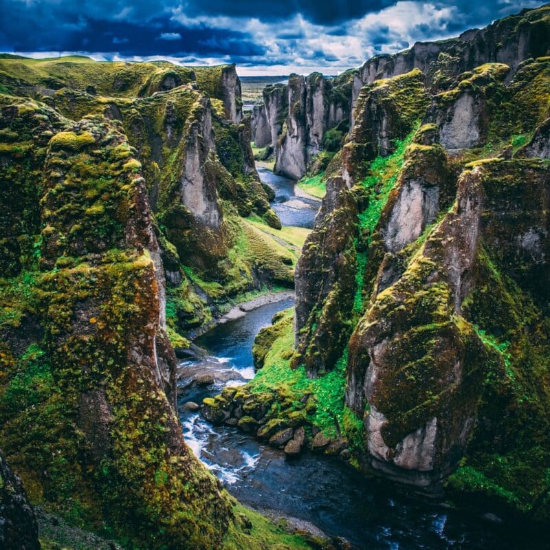 A dramatic Icelandic canyon with steep, moss-covered rock formations and a winding river flowing through the valley under a cloudy sky