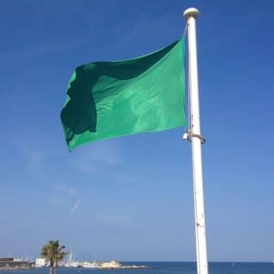 The old Libyan flag flying on a white flagpole near a coastal area with palm trees and boats in the background