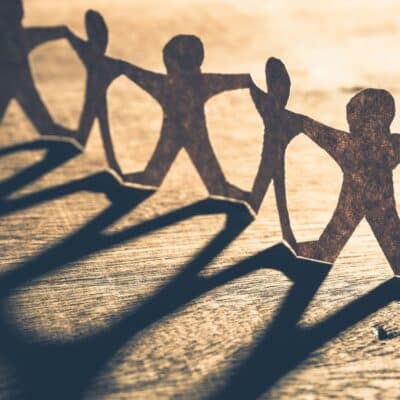 Paper cutouts of human figures holding hands, casting long shadows on a wooden surface