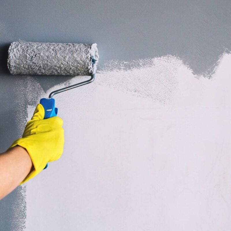 A person wearing yellow gloves uses a roller brush to paint a wall white