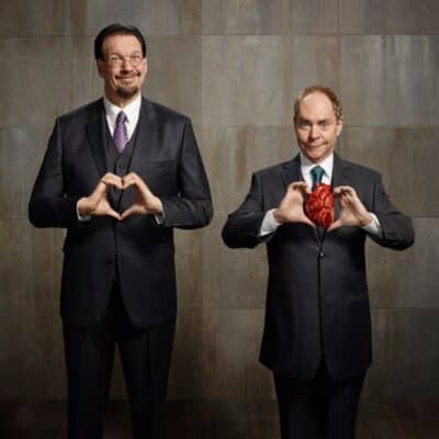 Penn and Teller in suits; Penn is making a heart shape with his hands, while Teller holds a realistic-looking heart