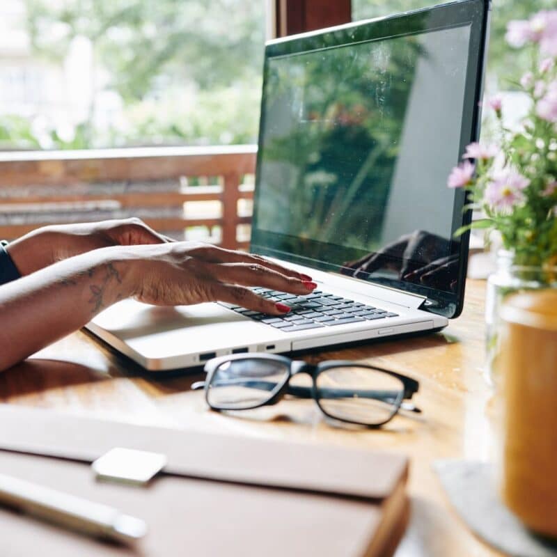 Hands typing on a laptop in a home office setting with a notebook, glasses, and flowers
