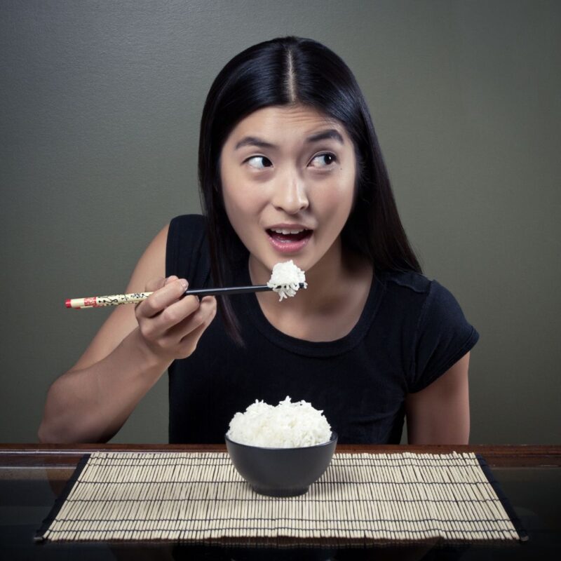 A woman holding chopsticks with a bite of white rice, sitting at a table with a black bowl full of rice on a bamboo mat