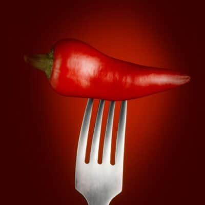 A single red chili pepper balanced on the prongs of a fork against a dark red background