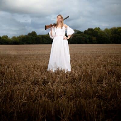 A woman in a white dress stands confidently in a field, holding a shotgun on her shoulder