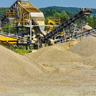 Large piles of industrial sand and gravel near a processing facility with conveyor belts and steel structures, set against a backdrop of trees and hills