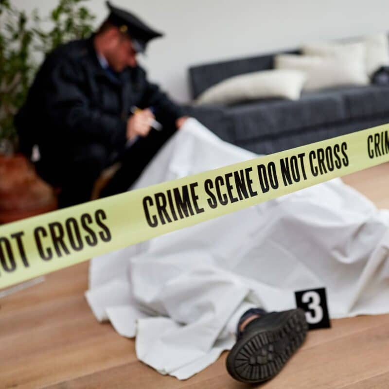 Police officer inspecting a covered body within a crime scene marked by yellow tape indoors