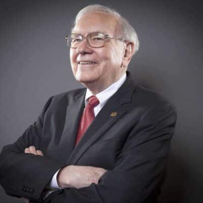 Warren Buffett wearing a dark suit with a red tie and smiling with his arms crossed