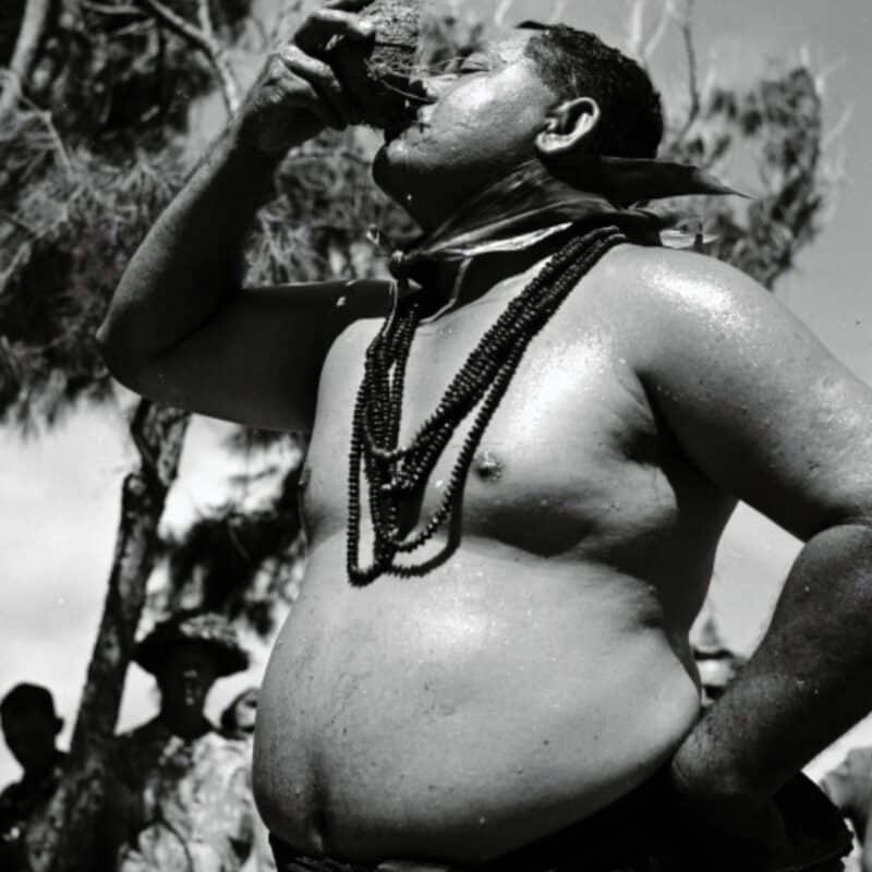 A shirtless man wearing beaded necklaces drinking from a coconut under a tree