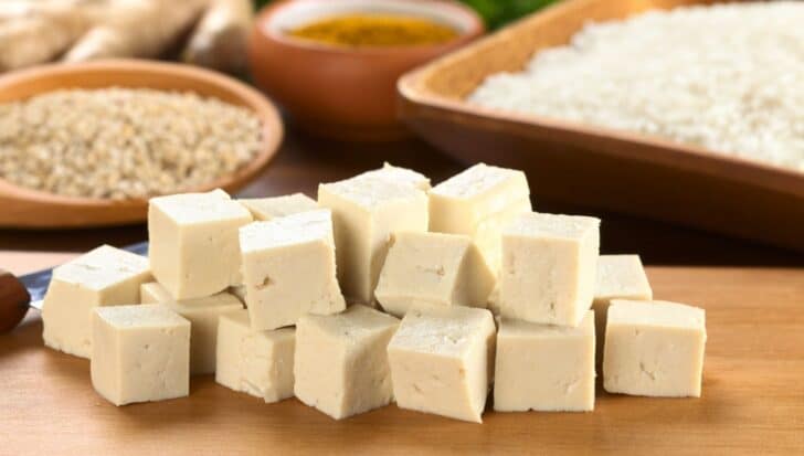 Cubes of tofu on a wooden surface with bowls of grains and spices in the background