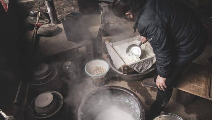 A person making tofu in a traditional setting with various cooking utensils and steaming pots visible