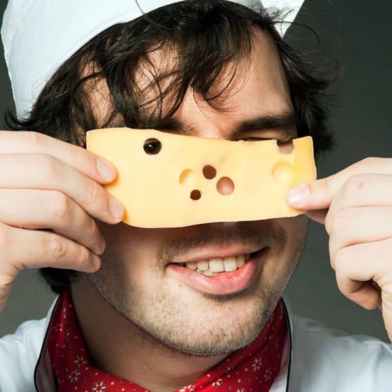 A smiling man in a white chef's hat holding a slice of Swiss cheese with holes up to his face