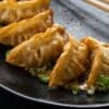 Fried dumplings arranged on a black plate with sliced green onions and sesame seeds, accompanied by wooden chopsticks