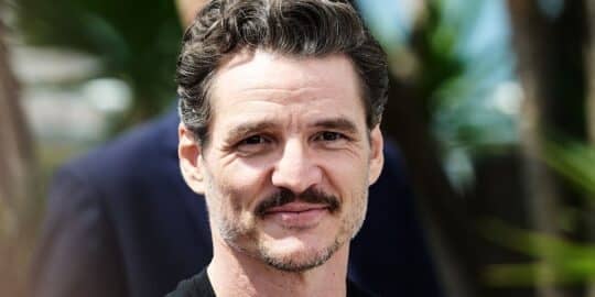 Pedro Pascal with short, wavy hair and a mustache smiling outdoors with blurred greenery in the background