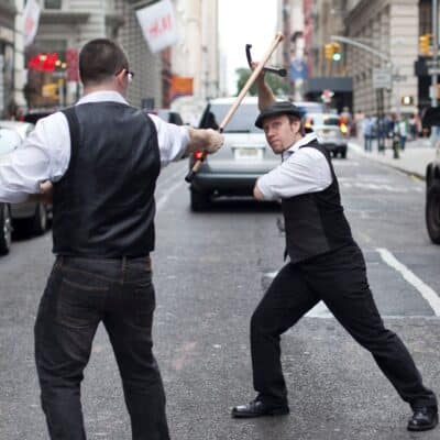 Two men in vintage vests spar with canes on a city street