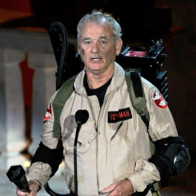 Bill Murray dressed in a Ghostbusters costume, holding a microphone