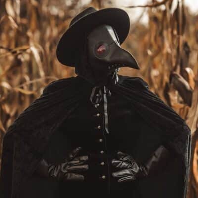 Person in black plague doctor costume standing in a dry cornfield