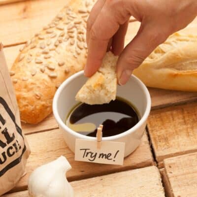 Hand dipping bread into olive oil and balsamic vinegar beside loaves and garlic