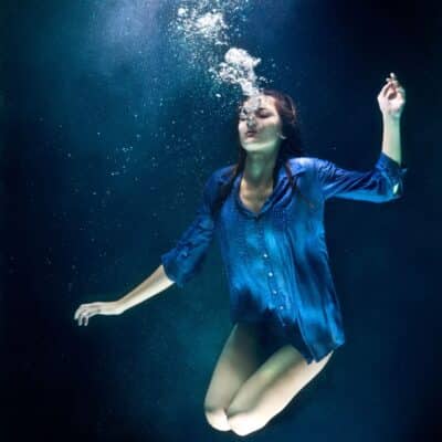 A woman in a blue shirt submerged underwater, exhaling bubbles