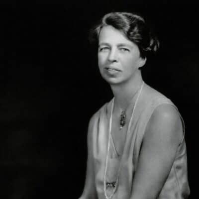 Eleanor Roosevelt wearing a sleeveless dress and long necklace, seated against a dark background