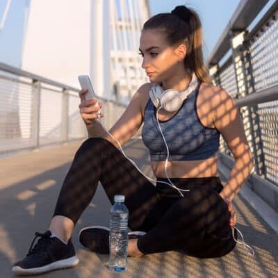 Woman in workout clothes sitting on bridge, looking at phone with headphones around neck