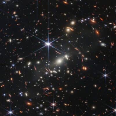 Dense field of distant galaxies, bright spiky stars, and orange lensing arcs