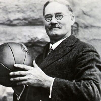 James Naismith wearing glasses and a suit holding a vintage basketball
