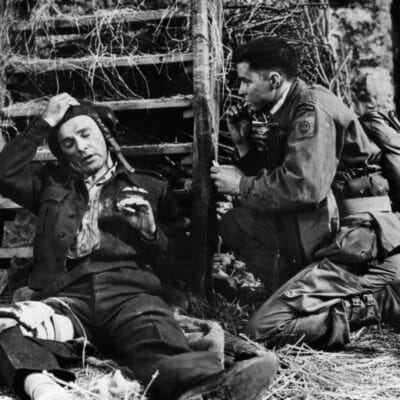 Two soldiers in WWII uniforms rest beside hay and wooden debris