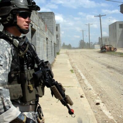 U.S. soldier in tactical gear stands near abandoned buildings and a burned vehicle