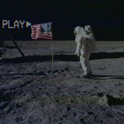 Buzz Aldrin stands beside American flag on the Moon with 'PLAY' text overlay