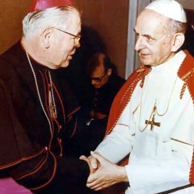 A bishop wearing a red zucchetto and black clerical attire with red accents, shaking hands with Pope Paul VI