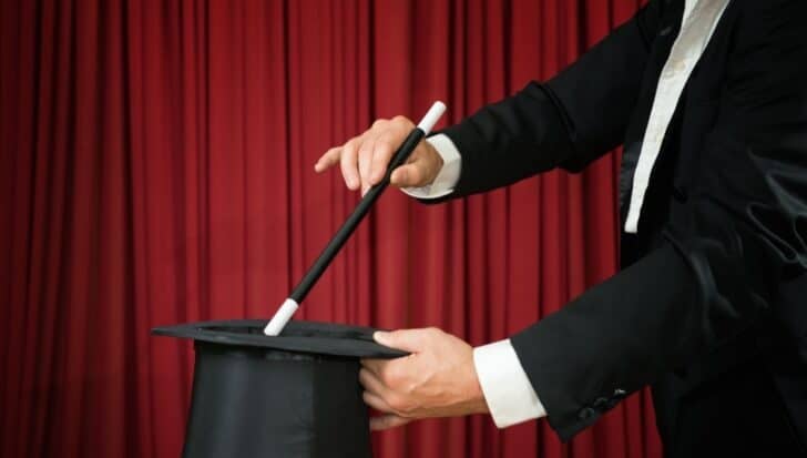 A magician in a suit holding a wand and top hat in front of a red curtain