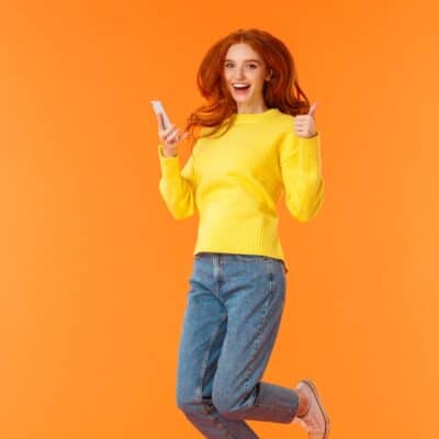 Smiling woman in yellow sweater giving thumbs up while holding smartphone