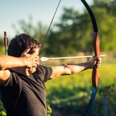 Man aiming a drawn bow and arrow outdoors in sunlight