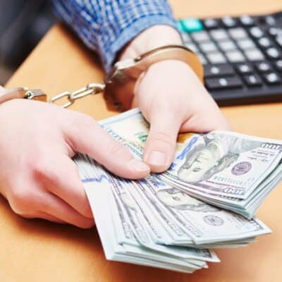Handcuffed person holding a stack of hundred-dollar bills near a keyboard