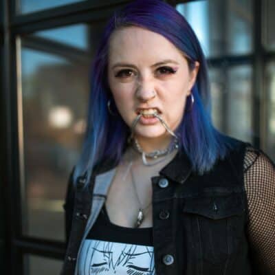 Woman with purple hair biting a chain, wearing hoop earrings and a graphic tank top