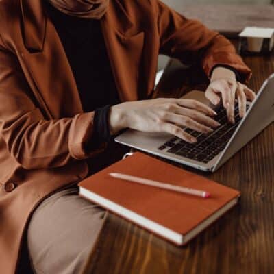 Person in a brown coat working on a laptop at a wooden table