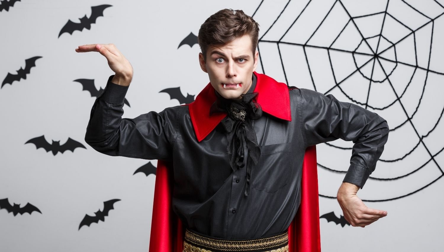 A man in a black suit and a red cape and vampire teeth in a silly pose.