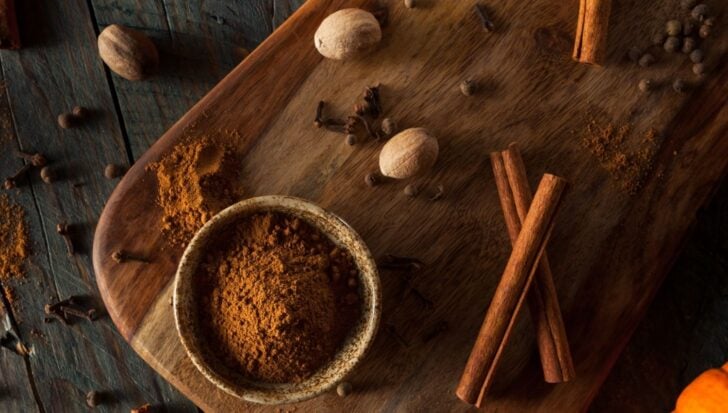 A wooden board with cinnamon sticks, nutmeg, cloves, and a bowl of pumpkin spice powder