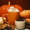A warm setting with an orange mug topped with whipped cream, surrounded by pumpkins, ginger, cinnamon sticks, cloves, and a small bowl of spices