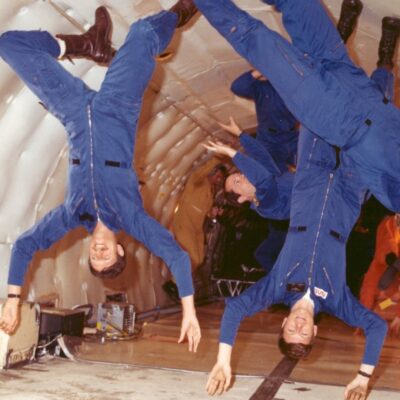 Astronauts in blue flight suits float upside down inside a microgravity training aircraft