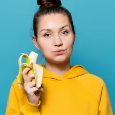 Woman in yellow hoodie holding a peeled banana against blue background