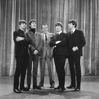 The Beatles pose with Ed Sullivan on a stage with curtains