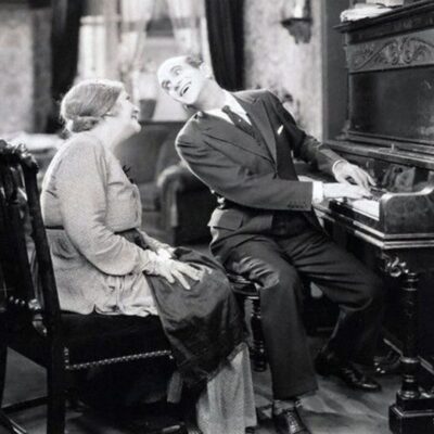 Man playing piano and singing beside an elderly woman in a vintage room