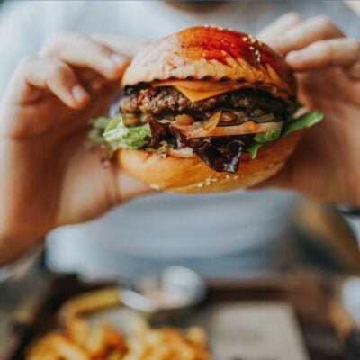 Person holding a large hamburger with lettuce, tomato, cheese, and onions