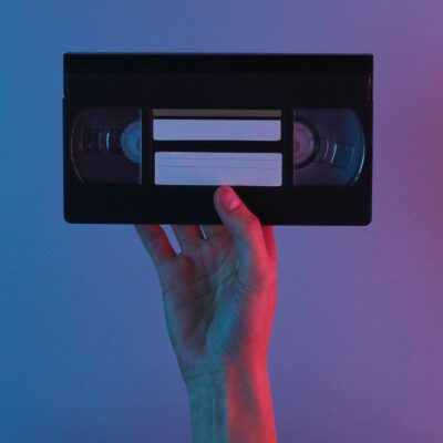 Hand holding a VHS tape against a pink and blue background