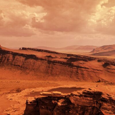 Red rocky landscape with layered cliffs under a cloudy sky, resembling Mars